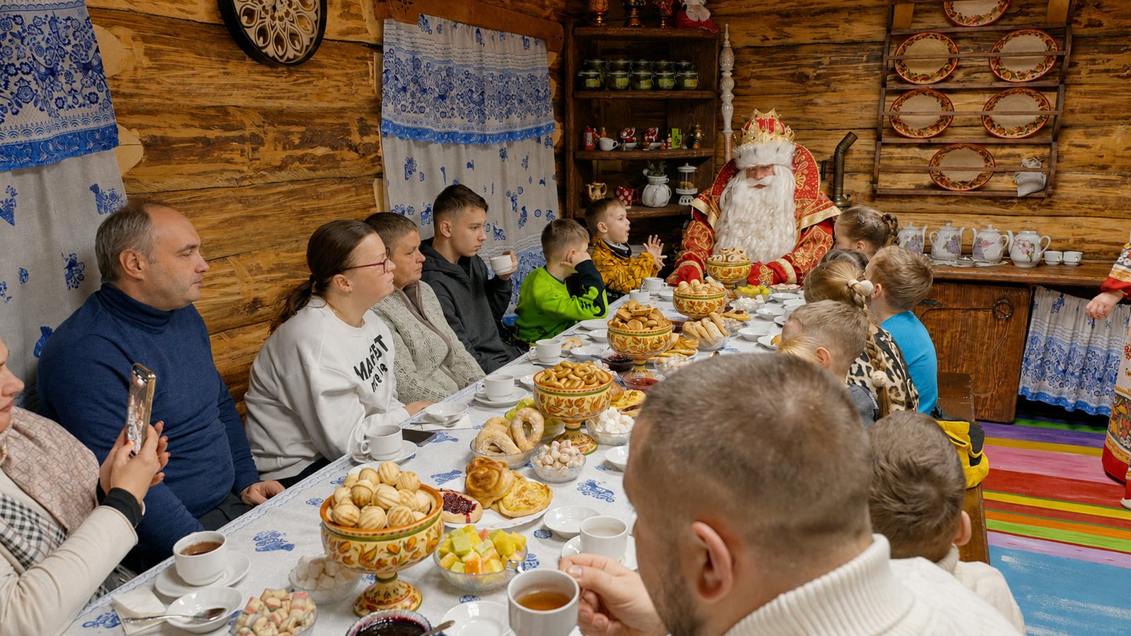 Большой накрытый стол с выпечкой, чашками и гостями; Дед Мороз виден в глубине комнаты.